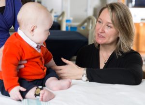 Baby on left sitting upright in red sweater facing researcher Robyn Stremler 