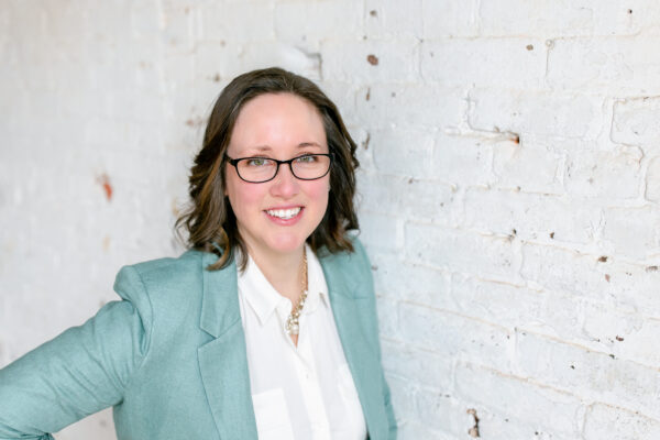 Profile of Amy Wright in teal blazer wearing glasses against white brick background