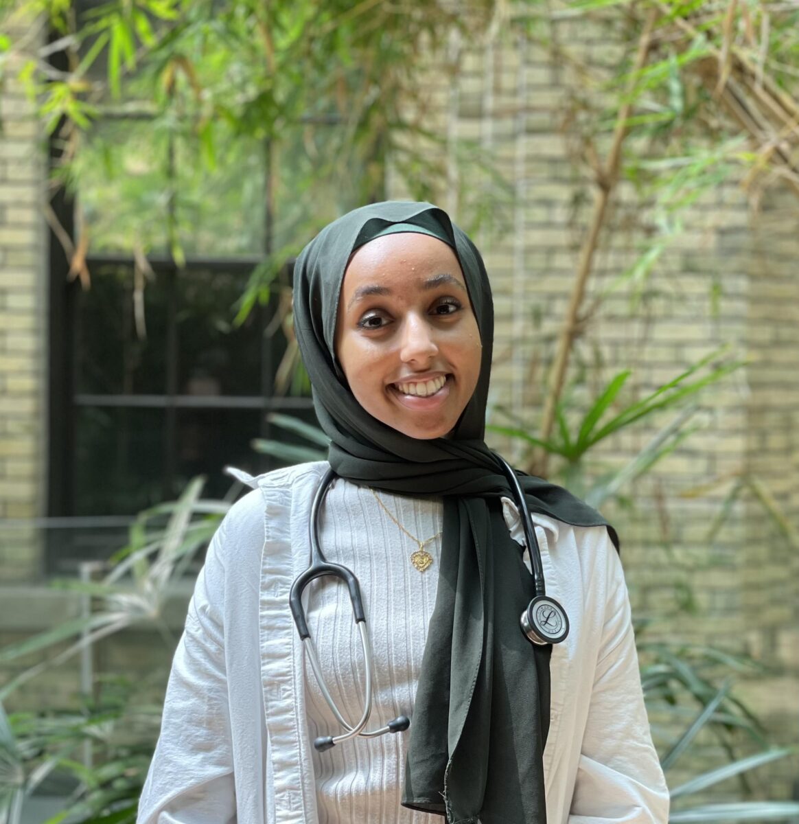 Image of woman in a hijab and wearing a stethoscope. Greenery and brick wall in the background